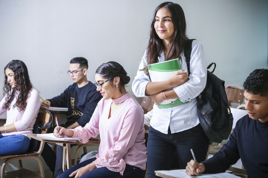 Students working at desks in a classroom, one standing with notebooks under her arm.